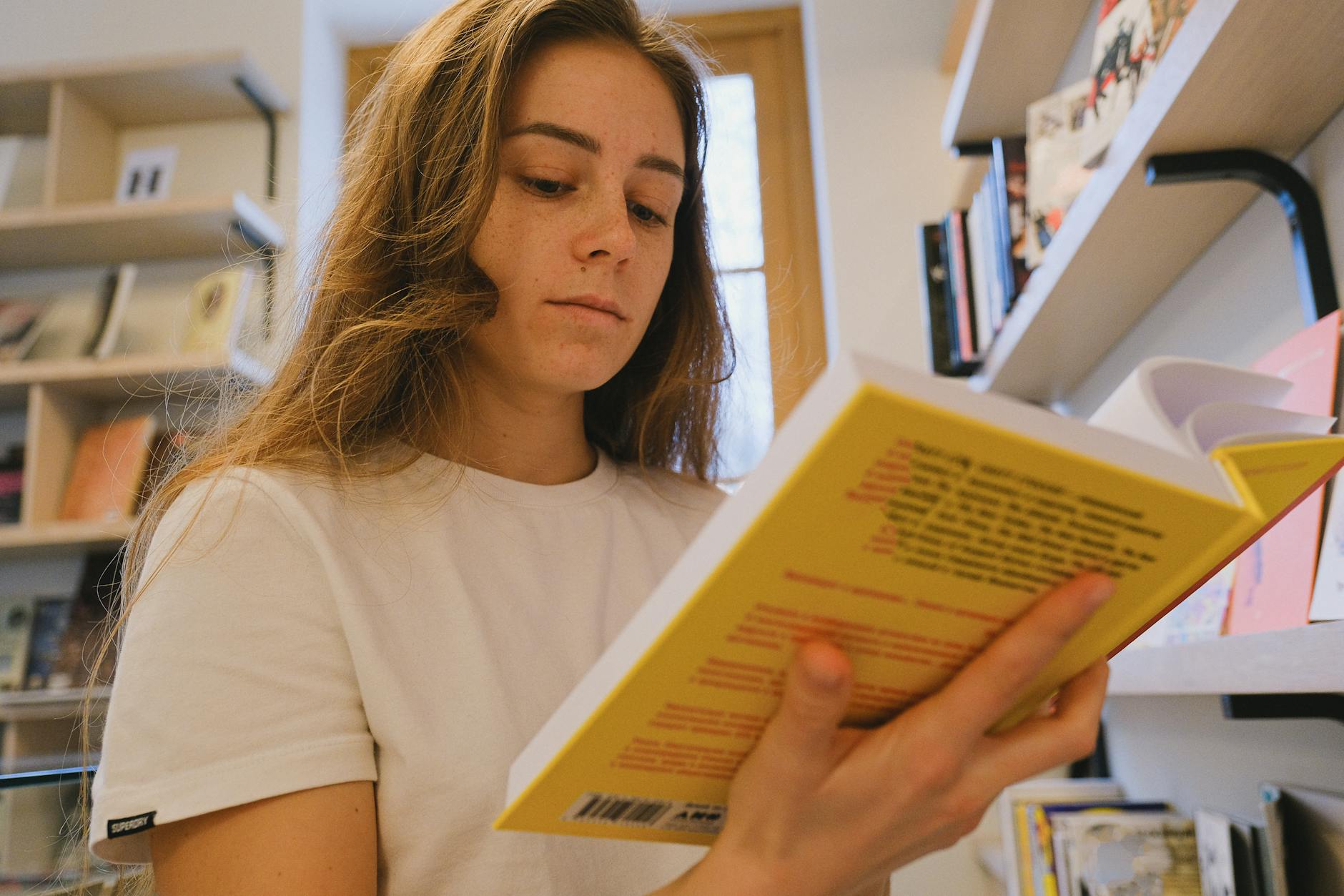 Młoda kobieta czyta żółtą książkę między regałami biblioteki Młoda kobieta czyta żółtą książkę między regałami biblioteki